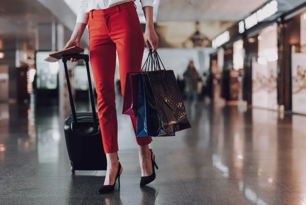 Woman, seen from the waist down, wearing high heels and carrying a rolling suitcase and shopping bags in an airport
